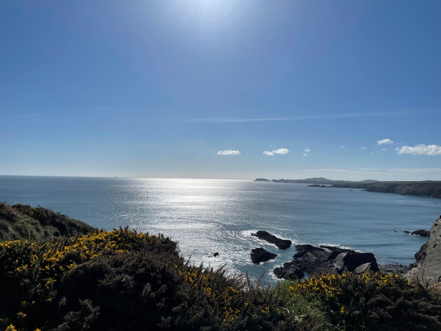 The sun reflecting off the open sea between Nine Wells and Solva, Penbrokeshire. The cloudless sky is bright blue and the sea is still, the sun is making a channel, reflecting off the water, in towards the coastline, breaking only around scatter rocks just off the shoreline which curves around in the distance on the right-hand side of the image
