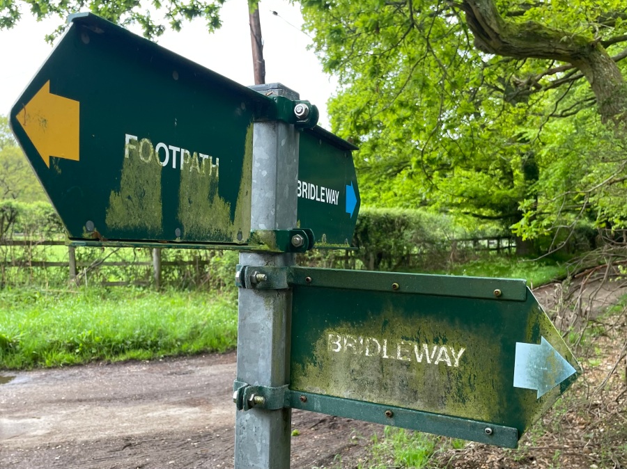 A three-way cross roads of faded green footpaths signs. To the left and right are footpaths and perpendicular to these is a bridle way