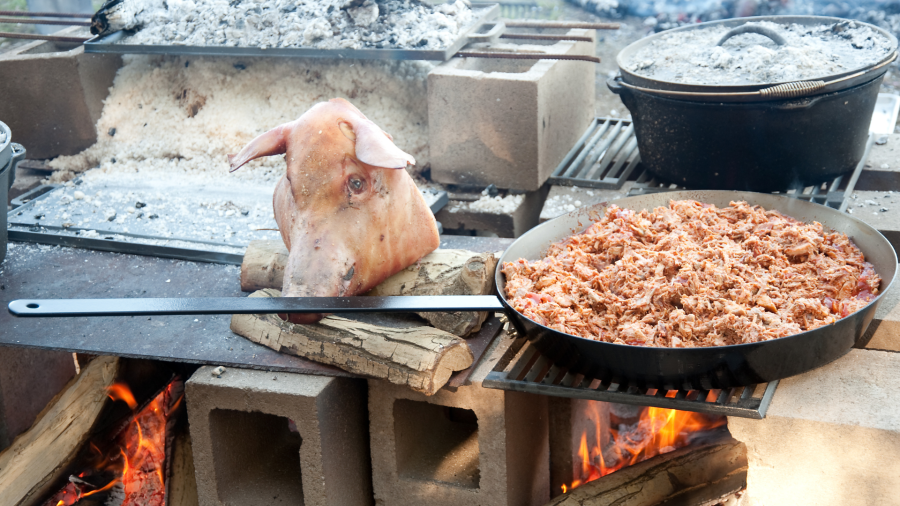 A large frying pan full of shredded meat on a homemade outdoor grill. The pan handle is being held in place by a stone pig’s head statue